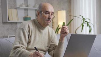 stressato e riflessivo vecchio uomo fare attività commerciale su computer. vecchio uomo riflessivo e preoccupato mentre fare il suo opera su il computer portatile a casa. video