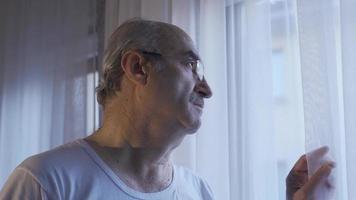 Unhappy old man looking out the window in the living room at home takes off his glasses. Pensive, unhappy old man thinking about loneliness, looking out the window. Cinematic old people scenes. video