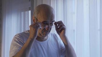 un pensativo antiguo hombre sentado solo a hogar es mirando fuera el ventana. preocupado antiguo hombre mira fuera el ventana y es considerado. video