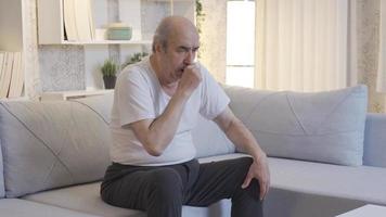 Old man sitting on sofa at home coughing and getting worse. A sick mature elderly man is coughing and is in critical health. video