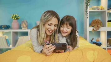 mamma och henne liten dotter är spelar på de telefon i säng på Hem. mor spelar med henne söt liten dotter i säng med henne telefon. söt mor och dotter. video