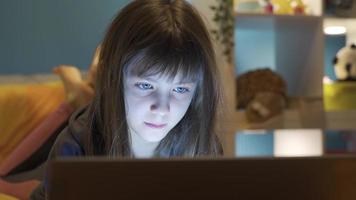 pequeno menina olhando às computador portátil dentro noite sala. feliz pequeno garota. a pequeno menina é navegando social meios de comunicação, mensagens de texto e gastos Tempo dentro frente do dela computador portátil às noite dentro dela sala. video