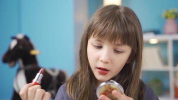 weinig meisje verven haar lippen met lippenstift. aan het doen verzinnen. grappig weinig meisje hebben pret aan het leren naar Doen bedenken in voorkant van klein spiegel in haar kamer. video