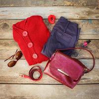 Women's autumn clothing and accessories red sweater, pants, handbag, beads, sunglasses, nail polish, hair band, belt on wooden background. photo