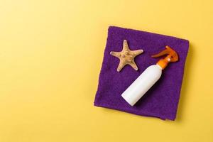 Beach flat lay accessories with copy space. Violet blue and white towel, seashells, staw sunhat and a bottle of sunblock on yellow background. Summer holiday concept photo