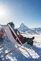 Young snowboarder cheering with a beer after skiing day in a bar or a cafe at the Zermatt ski resort in Swiss Alps with a fabulous view of the Matterhorn. Winter vacation concept. photo