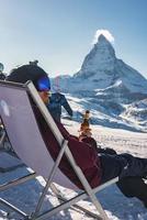 Young snowboarder cheering with a beer after skiing day in a bar or a cafe at the Zermatt ski resort in Swiss Alps with a fabulous view of the Matterhorn. Winter vacation concept. photo