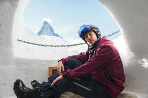 Portrait of smiling tourist snowboarder sitting inside an iglu dorf with a view on the famous snowcapped Matterhorn mountain. Relaxing in Swiss Alps, Zermatt ski resort. photo