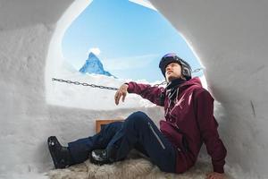 Portrait of smiling tourist snowboarder sitting inside an iglu dorf with a view on the famous snowcapped Matterhorn mountain. Relaxing in Swiss Alps, Zermatt ski resort. photo