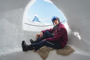 Portrait of smiling tourist snowboarder sitting inside an iglu dorf with a view on the famous snowcapped Matterhorn mountain. Relaxing in Swiss Alps, Zermatt ski resort. photo