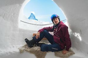 Portrait of smiling tourist snowboarder sitting inside an iglu dorf with a view on the famous snowcapped Matterhorn mountain. Relaxing in Swiss Alps, Zermatt ski resort. photo