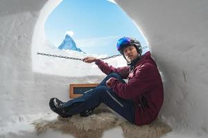 Portrait of smiling tourist snowboarder sitting inside an iglu dorf with a view on the famous snowcapped Matterhorn mountain. Relaxing in Swiss Alps, Zermatt ski resort. photo