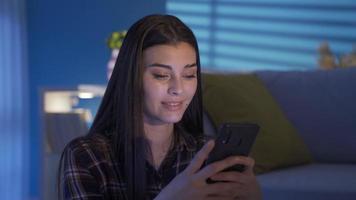 contento joven mujer sentado en el piso por el sofá mirando a el teléfono con un sonrisa. enviar mensajes de texto joven mujer sonriente, disfrutando utilizando móvil aplicaciones para compras, teniendo divertido, chateando en social medios de comunicación. video