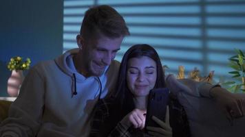 Happy couple laughing looking at smartphone screen at night at home. Happy young couple sitting on sofa at home at night looking at funny social media posts on the phone. video