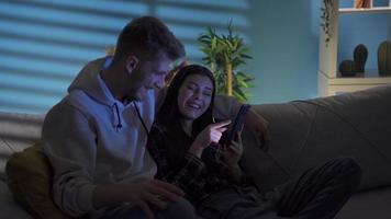 Smiling positive happy couple looking at funny videos on social media on smartphone. Happy young couple sitting on sofa at home at night looking at funny social media posts on the phone.