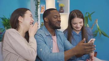 Multiethnic young people looking at phone at home chatting and laughing. Young people looking at the phone laugh out loud. Asian woman, European woman and African man. video