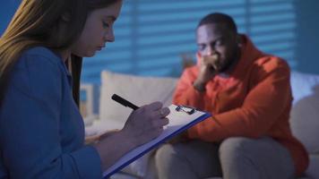 Female psychologist listening to black man with psychological problems and making notes on paper. video