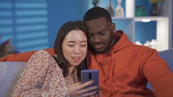 Happy multiracial couple sitting on sofa at home chatting, looking at phone and laughing. Asian beautiful woman and handsome black man sitting on sofa at home, looking at phone. video