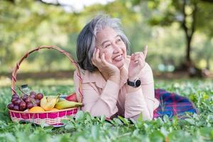 Happy Asian old senior woman and wear a health watch and lying on the picnic mat in park and basket of fruit besides. Concept of happy elderly woman after retirement and good health photo