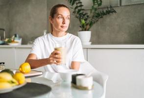 Young slim woman in white t-shirt and blue jeans drinking fruit smoothie healthy food in kitchen at home photo