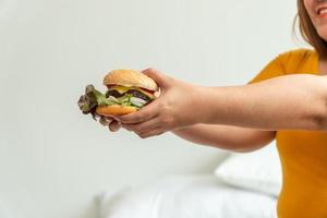 Hungry overweight woman smiling and holding hamburger and sitting in the bedroom, her very happy and enjoy to eat fast food. Concept of binge eating disorder BED photo