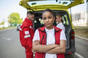 joven hembra africano americano paramédico en pie posterior de el ambulancia. paramédicos por el ambulancia. dos paramédicos tomando fuera camilla desde ambulancia foto