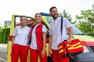 paramédicos y médico en pie en el lado ambulancia. médico es que lleva un médico trauma bolsa. grupo de Tres paramédicos en pie en frente de ambulancia con sonrisa. foto