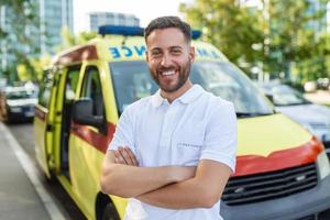 joven hombre , un paramédico, en pie a el posterior de un ambulancia, por el abierto puertas él es mirando a el cámara con un confidente expresión, sonriente foto