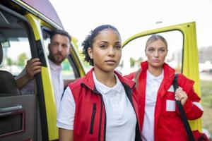 paramédicos y médico en pie en el lado ambulancia. médico es que lleva un médico trauma bolsa. grupo de Tres paramédicos en pie en frente de ambulancia con sonrisa. foto