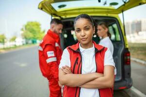 joven hembra africano americano paramédico en pie posterior de el ambulancia. paramédicos por el ambulancia. dos paramédicos tomando fuera camilla desde ambulancia foto