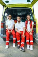 multiétnico grupo de paramédicos en pie a el lado de un ambulancia con abierto puertas su compañero de trabajo que lleva un médico trauma bolsa. ellos son sonriente a el cámara. foto