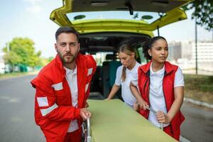 joven paramédicos Moviente ambulancia camilla desde coche en un apuro. paramédicos en uniforme tomando camilla fuera el ambulancia coche foto