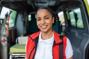 Young woman , a paramedic, standing at the rear of an ambulance, by the open doors. She is looking at the camera with a confident expression, smiling, carrying a medical trauma bag on her shoulder. photo