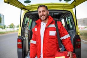 Young man , a paramedic, standing at the rear of an ambulance, by the open doors. He is looking at the camera with a confident expression, smiling, carrying a medical trauma bag on his shoulder. photo