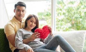 contento Pareja sentado en el sofá y siendo un hombre abrazando su Novia con amor en el vivo habitación y sonrisa y participación un rojo corazón. concepto de romántico en enamorado día. propuesta y matrimonio foto