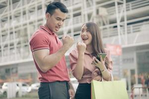 Happy young couple of shoppers walking in the shopping street towards and holding colorful shopping bags in hand and use a smartphone for check promotion. Concept of sale and Black Friday shopping photo