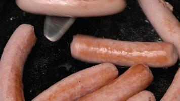Cooking and stirring sausages fried in black pan with oil. Close-up view of hands chef stirs sausages with metal spatula video