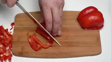 dichtbij omhoog visie van Kaukasisch mannetje chef hakken rood klok peper met groot mes in klein plakjes Aan houten keuken snijdend bord. snijden zoet peper, werk werkwijze van Koken groenten voor salade video