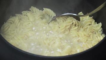 Cooking and stirring pasta in pan. Cook spaghetti stirs with metal spoon while traditional Italian cuisine cooking boiled in boiling water in black saucepan. Close-up view video