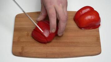 Caucasian male cook cook red capsicum with sharp knife into small slices on wooden kitchen cutting board. Close-up view of slicing sweet pepper, work process of cooking vegetables in kitchen video