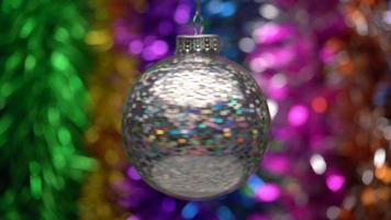 Beautiful multicolored Christmas ball hanging and spinning on blurry colored bokeh background of glowing tinsel, holiday lights. Close up of single holiday toy. Soft and selective focus on foreground video