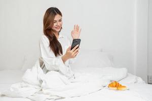 mujer en blanco bata de noche despertar arriba en fin de semana Mañana descansando y relajante jugando con ordenador portátil móvil teléfono comiendo un pan y Bebiendo té en vaso dentro blanco dormitorio. Mañana vacaciones concepto. foto