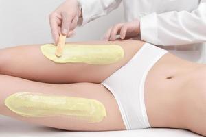 Beautician in gloves applying green hot wax on slim woman legs using spatula while woman lying photo