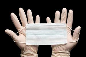 Doctor holds medical face mask in two hands in white gloves isolated on black background. Concept coronavirus quarantine photo