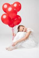 Romantic girl holding lot of red balloons, put head on knees, sitting on white background