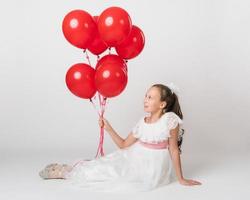 Beautiful girl dressed in long white dress holding lot of red balloons in hand, looking up at balls photo