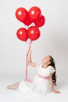 Smiling girl dressed in long white dress holding lot of party red balloons in hand, looking up at balls