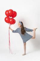 Playful girl holding large bunch of red balloons in hand, standing on one leg, looking at balloons