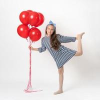 Active girl posing, holding bunch of red balloons in hand, standing on one leg on white background photo