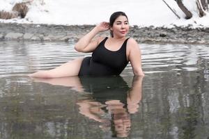 Curvy plus size young model in black bathing suit lying in geothermal water in outdoors pool at spa photo